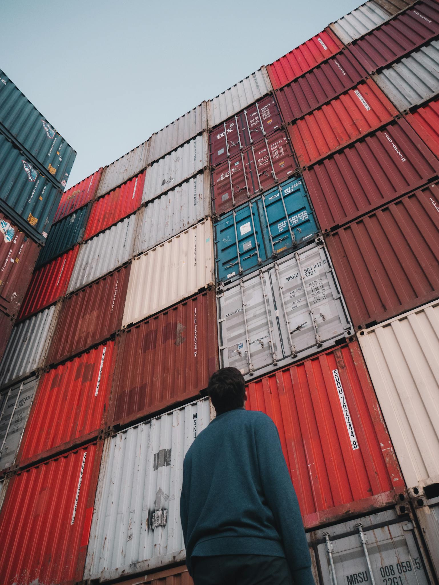 Man in blue sweater looking at high stacked shipping containers at a port.